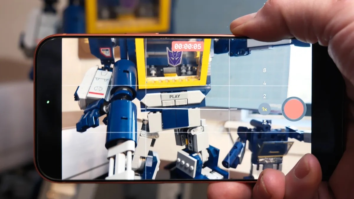 من موقع فون إسلام: شخص يستخدم الآي شخص يستخدم الآي شخص للتحكم بالكاميرا وتسجيل فيديو لدمية روبوت زرقاء وصفراء تظهر في الغرفة وعلى شاشة الهاتف أيضاً.