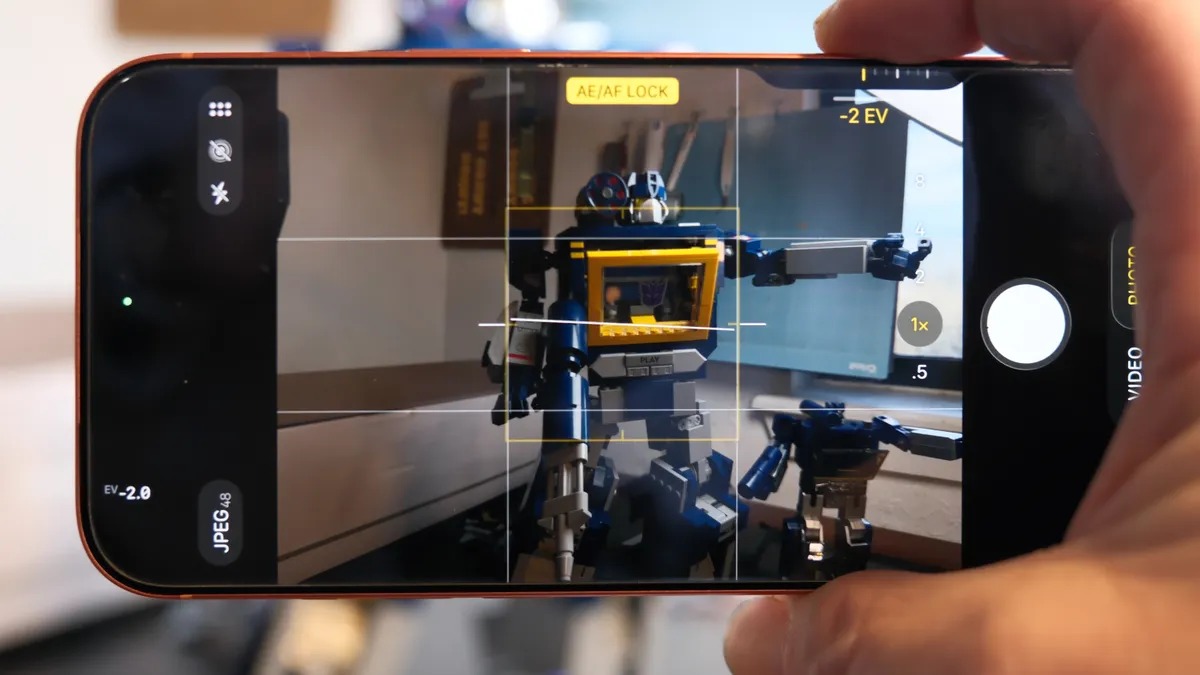من موقع فون إسلام: تعرض شاشة كاميرا الهاتف الذكي صورة يتم التقاطها لمجسم لعبة روبوت باللونين الأزرق والأصفر، مع مزايا التحكم بالكاميرا وخصائص الضبط في الآي-فون مثل التركيز والتعريض على الشاشة.