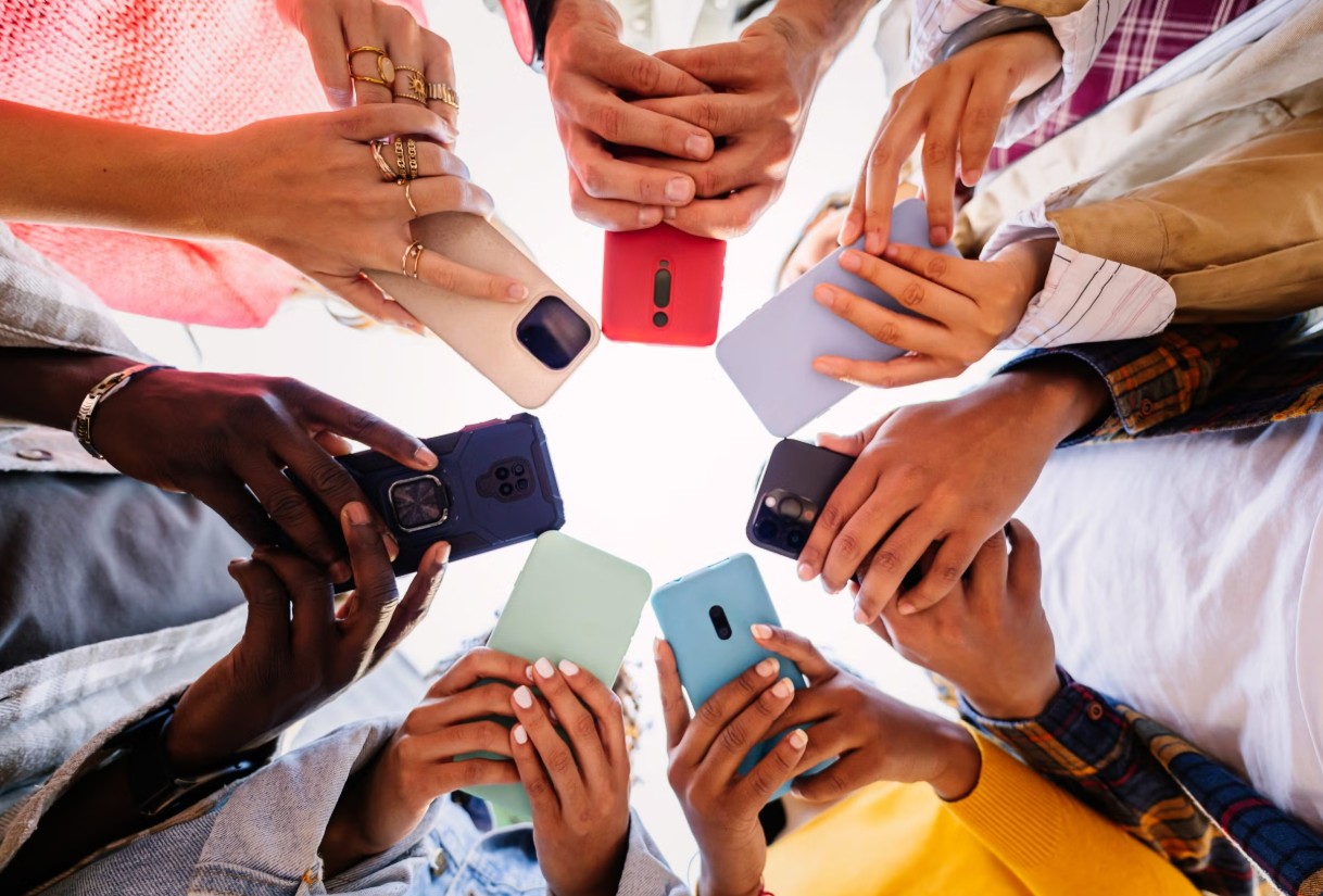 من موقع فون إسلام: مجموعة من الأشخاص يقفون في دائرة يحملون هواتف ذكية ذات أغلفة ملونة، ويشكلون حلقة بأيديهم وأجهزتهم التي ينظر إليها من الأسفل - وهو مشهد يذكرنا بالابتكار الذي يناصره تيم كوك.