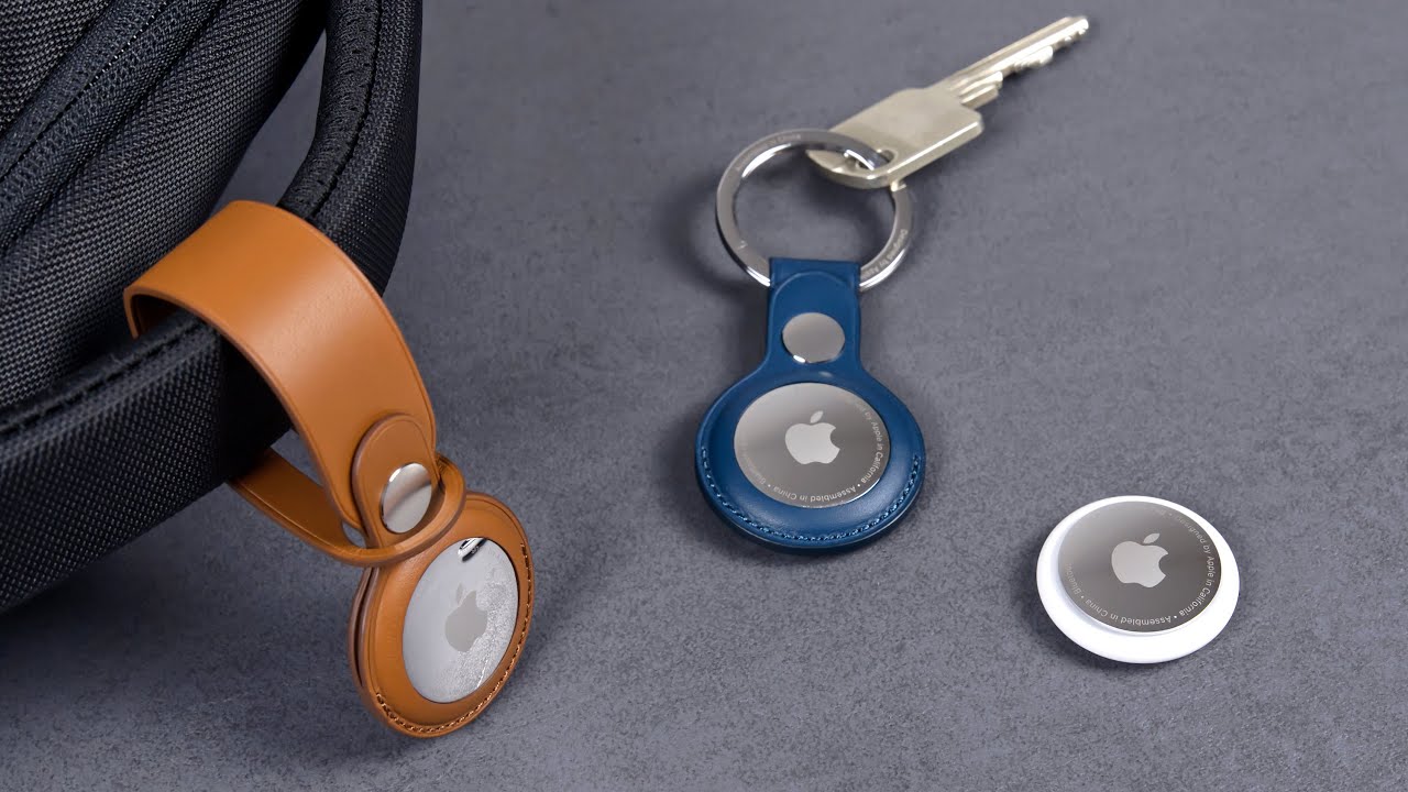 من موقع فون إسلام: علامات AirTags من Apple AirTags المعروضة: واحدة مثبتة بحلقة جلدية بنية اللون على حقيبة، وواحدة في حلقة مفاتيح زرقاء مع مفاتيح، وواحدة غير مثبتة على سطح رمادي - مثالية لتتبع أغراضك باستخدام ميزة AirTag.