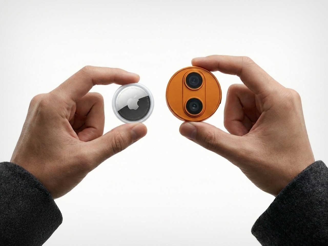 من موقع فون إسلام: تحمل كلتا اليدين جهازًا صغيرًا: تحمل اليد اليسرى جهاز AirTag من Apple، بينما تحمل اليد اليمنى جهازًا دائريًا برتقالي اللون دبوس الذكاء الاصطناعي مع عدستين للكاميرا.