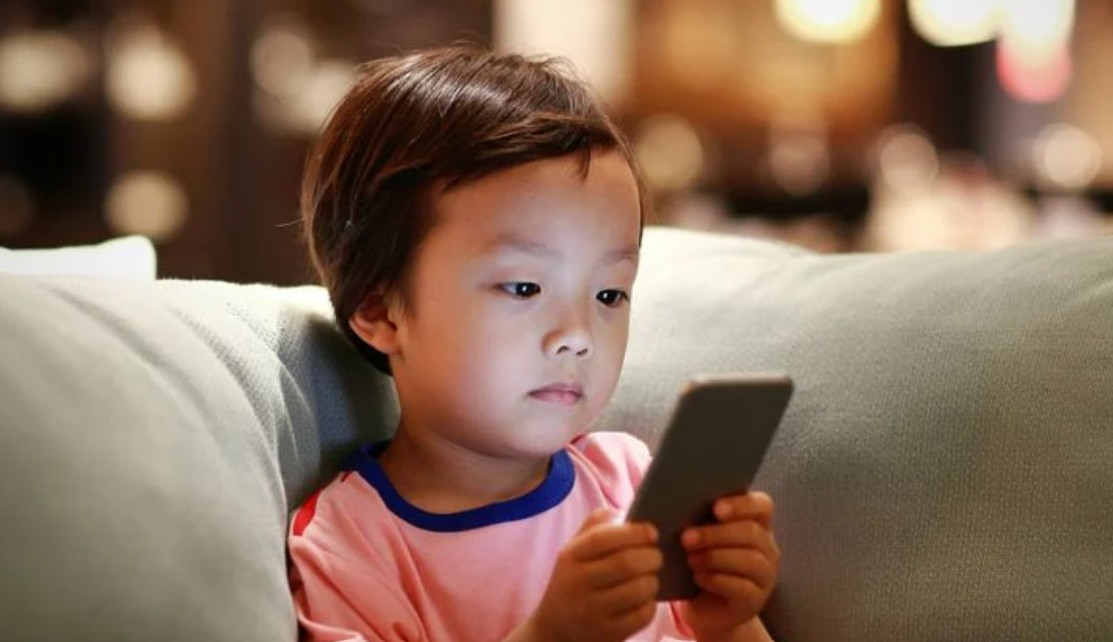 من موقع فون إسلام: طفل صغير يجلس على أريكة ويحمل هاتفًا ذكيًا وينظر إليه بتركيز شديد، ربما يكون مستوحى من ابتكارات تيم كوك.