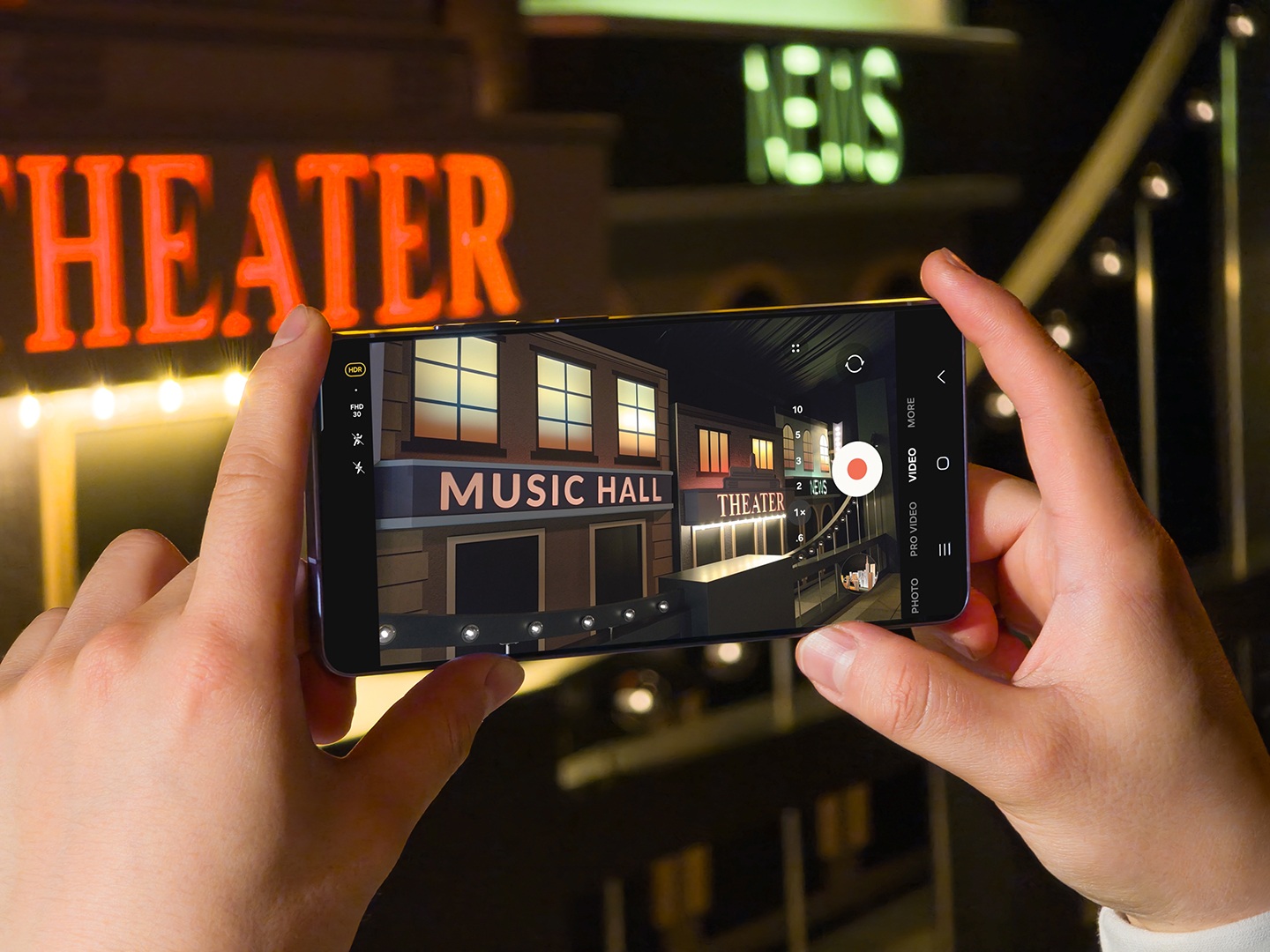 من موقع فون إسلام: شخص يحمل هاتفًا ذكيًا يصور مسرحًا مضاءً بشكل ساطع ولافتة قاعة موسيقى ليلاً - ملتقطًا الأجواء النابضة بالحياة التي تذكرنا بأحداث مؤتمر سامسونج.