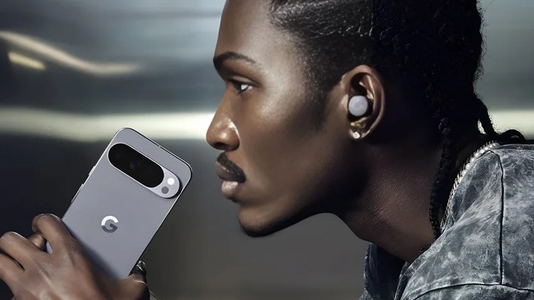 من موقع فون إسلام: شخص يرتدي سماعة أذن لاسلكية يحمل هاتفًا ذكيًا رمادي اللون من Google Pixel، يتميز بوحدة كاميرا خلفية كبيرة مزودة بثلاث عدسات، تذكرنا بالتصميم الذي يشاع عن الهاتف الذكي آي ئي ¬17 برو.