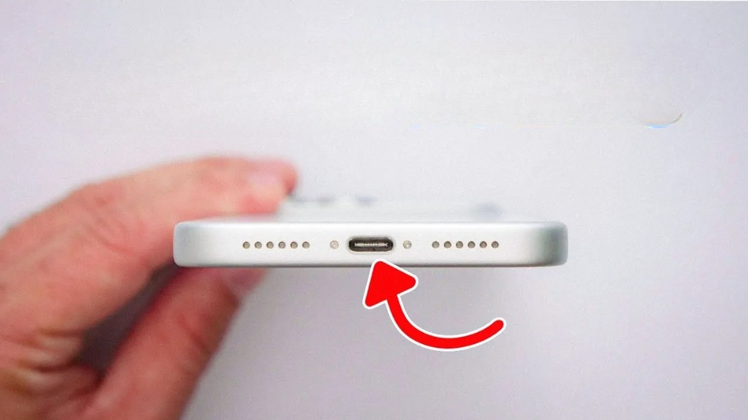 من موقع فون إسلام: يد تحمل هاتفًا ذكيًا يظهر على حافته السفلية منفذ شحن USB-C وشبكتي مكبر صوت وسهم أحمر يشير إلى المنفذ.