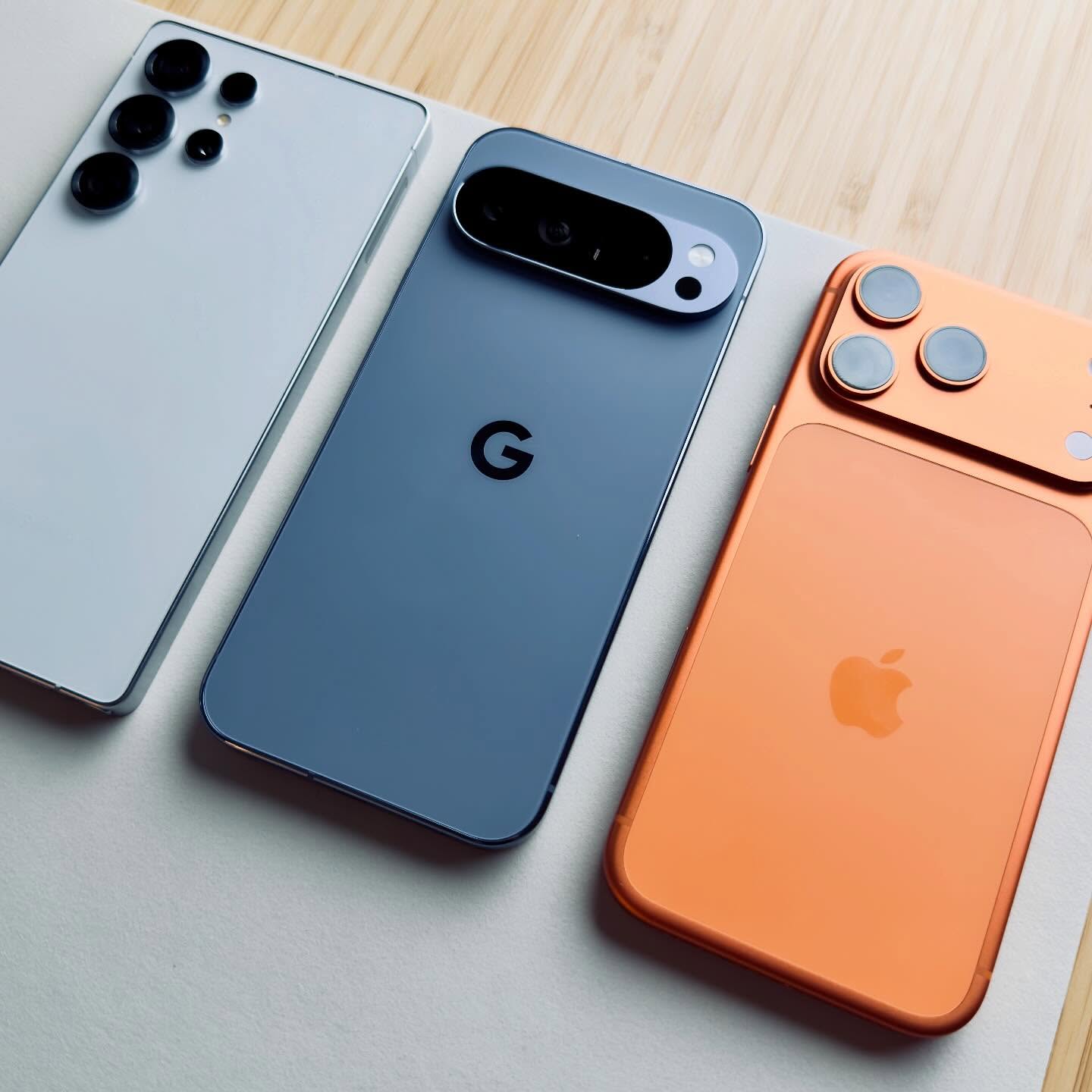 من موقع فون إسلام: ثلاثة هواتف ذكية - واحد أبيض والآخر رمادي مع شعار "G" والآخر برتقالي مع شعار آبل ربما إصدار آيفون 17 برو - موضوعة جنبًا إلى جنب على سطح فاتح اللون.