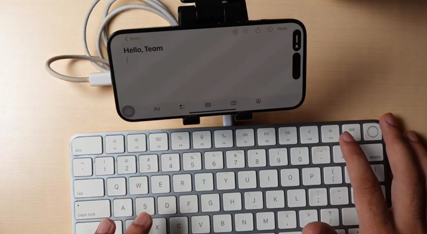 من موقع فون إسلام: يقوم شخص بكتابة "مرحباً أيها الفريق" على لوحة مفاتيح خارجية متصلة عبر USB-C بهاتف ذكي يعمل بنظام iOS 26، وهو مثبت على حامل فوق لوحة المفاتيح.