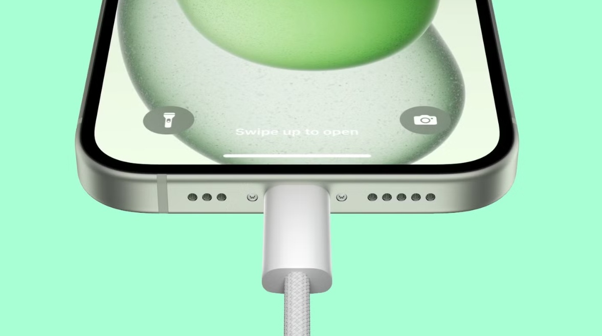 من موقع فون إسلام: لقطة مقرّبة لهاتف ذكي مزوّد بكابل شحن فاتح اللون موصول بكابل USB-C، تظهر فيه شاشة قفل iOS 26 وأيقونات للمصباح اليدوي والكاميرا.