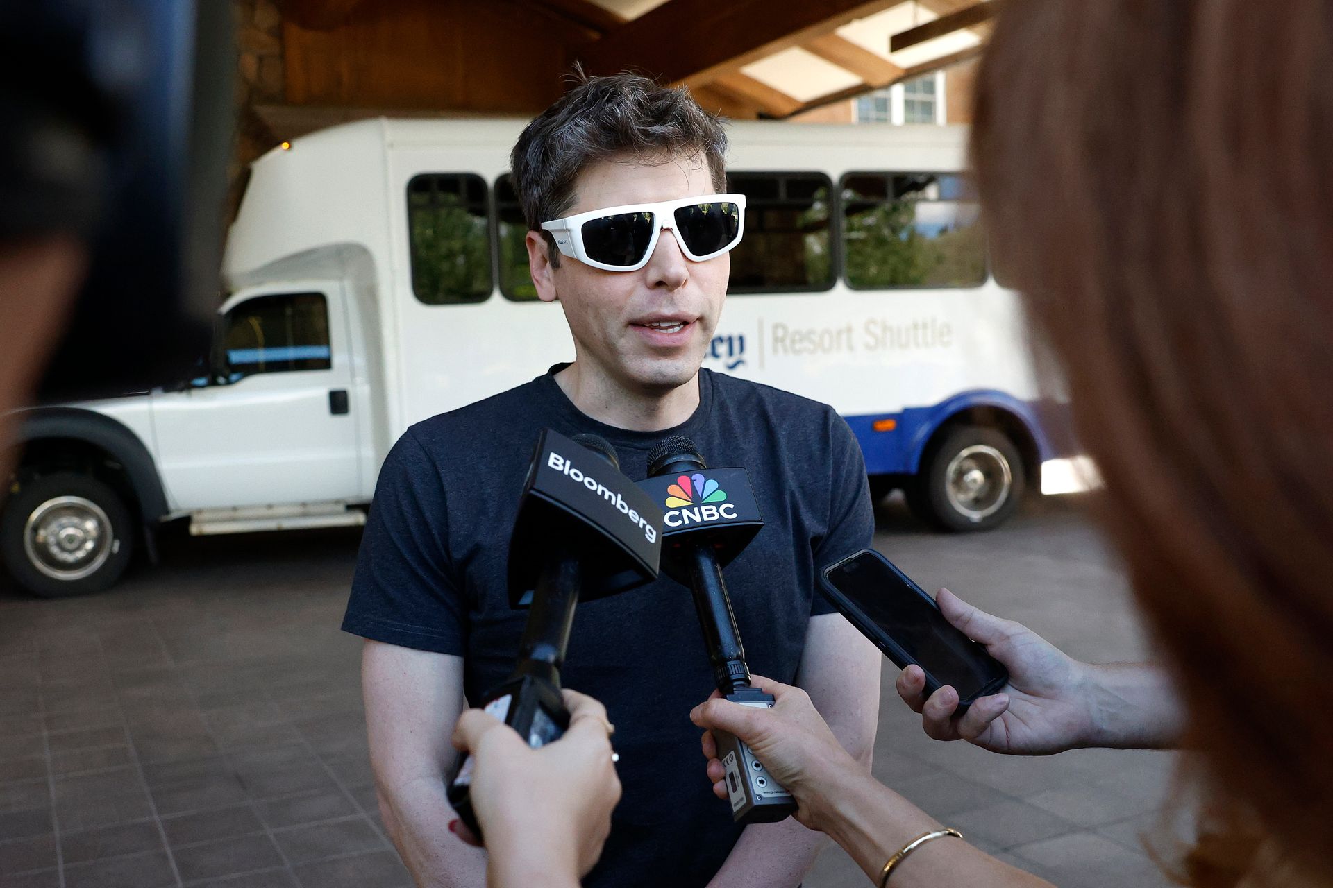 من موقع فون إسلام: رجل يرتدي نظارات شمسية وقميصاً أسود يتحدث إلى المراسلين الذين يحملون ميكروفونات بلومبرج وشبكة CNBC حول أخبار التكنولوجيا، وفي الخلفية حافلة بيضاء.
