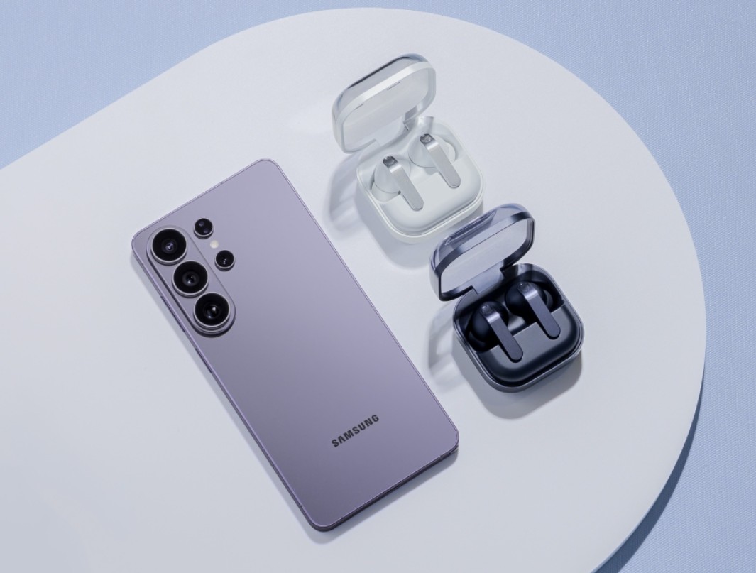 من موقع فون إسلام: تم وضع هاتف ذكي من سامسونج بلون الخزامى بجانب زوجين من سماعات الأذن اللاسلكية في علب شحن بيضاء وسوداء على سطح أبيض وأزرق فاتح، مستوحى من أحدث تصاميم مؤتمر سامسونج.