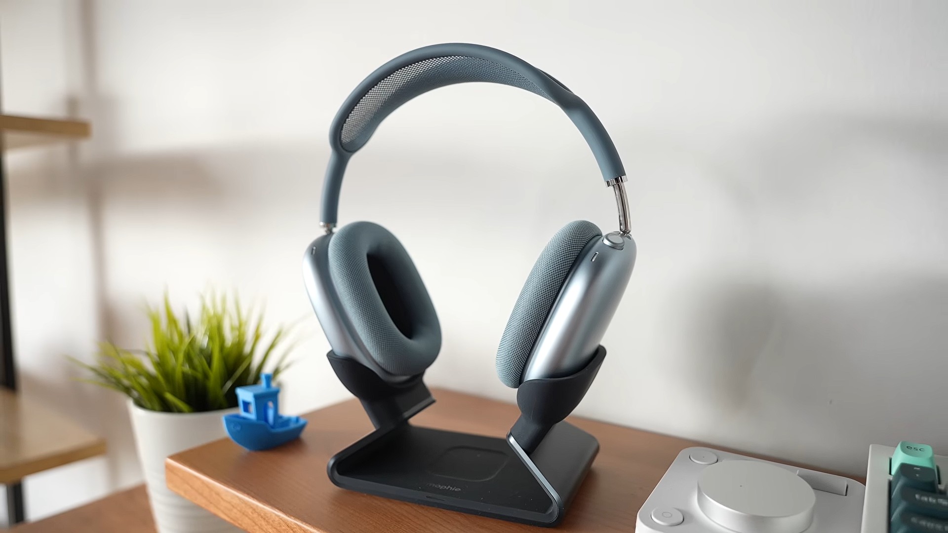 من موقع فون إسلام: زوج من سماعة AirPods Max 2 من سماعة AirPods Max 2 على حامل فوق مكتب خشبي، وبجواره نبتة صغيرة وأجهزة إلكترونية.