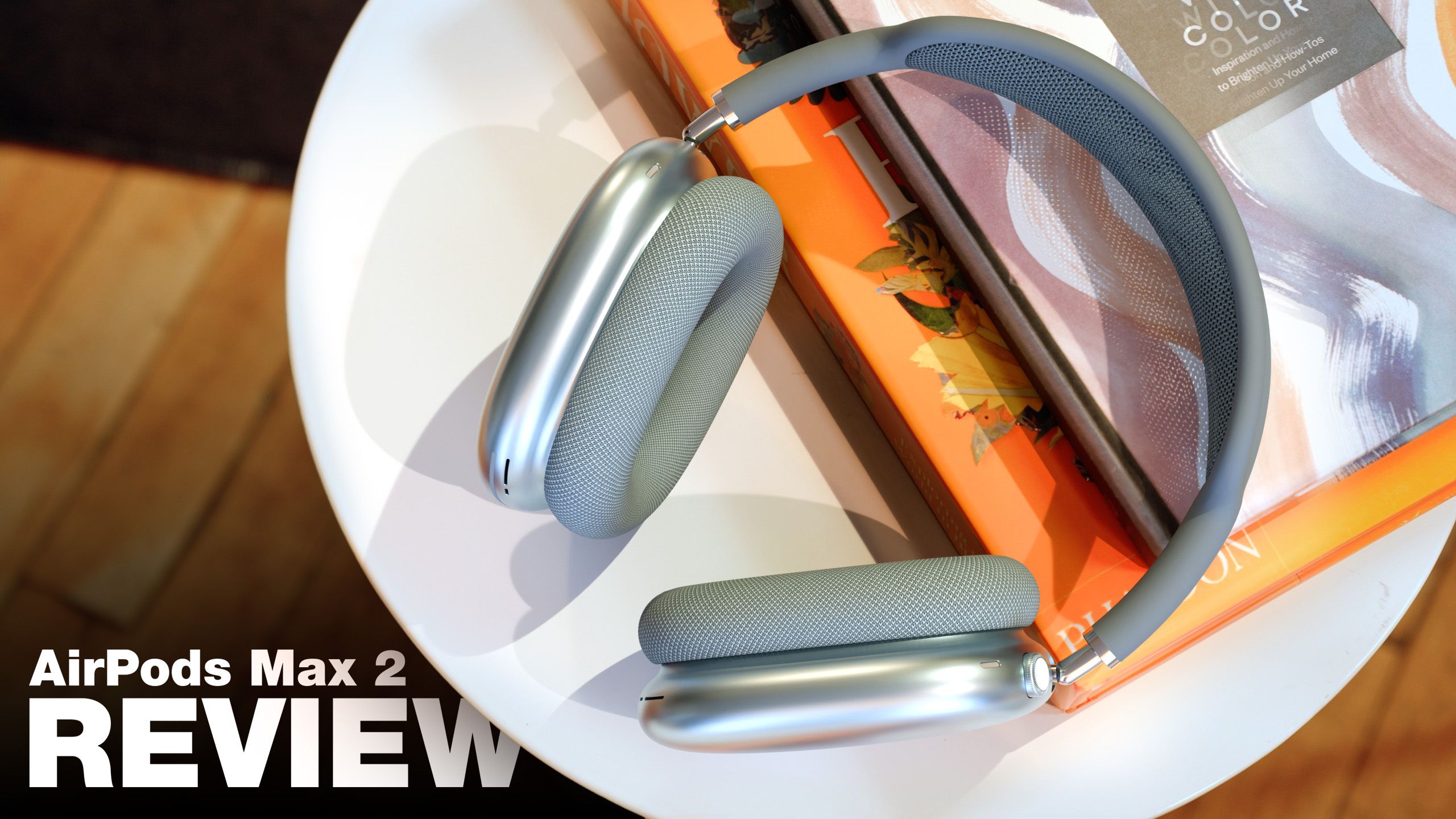 من موقع فون إسلام: زوج من سماعات الرأس الفضية التي توضع فوق الأذنين على طاولة بيضاء بجانب كتب ملونة، مع نص "AirPods Max 2 REVIEW" في الزاوية.