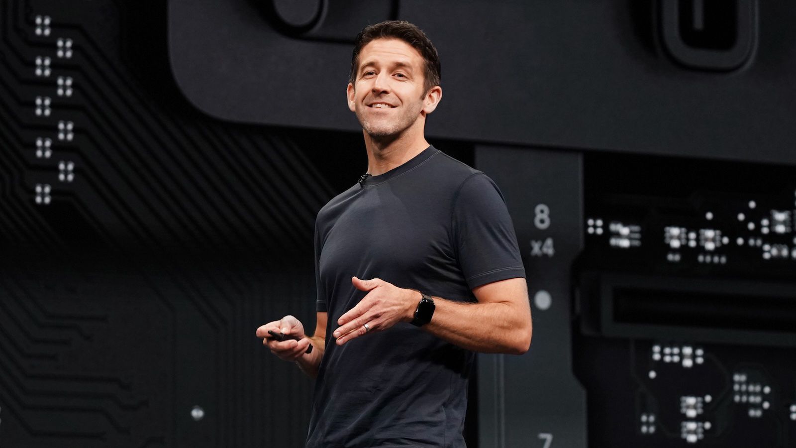 من موقع فون إسلام: رجل يرتدي قميصاً داكن اللون يقدم عرضاً على خشبة المسرح، ممسكاً بجهاز صغير، مع رسم لوحة دوائر كهربائية في الخلفية.