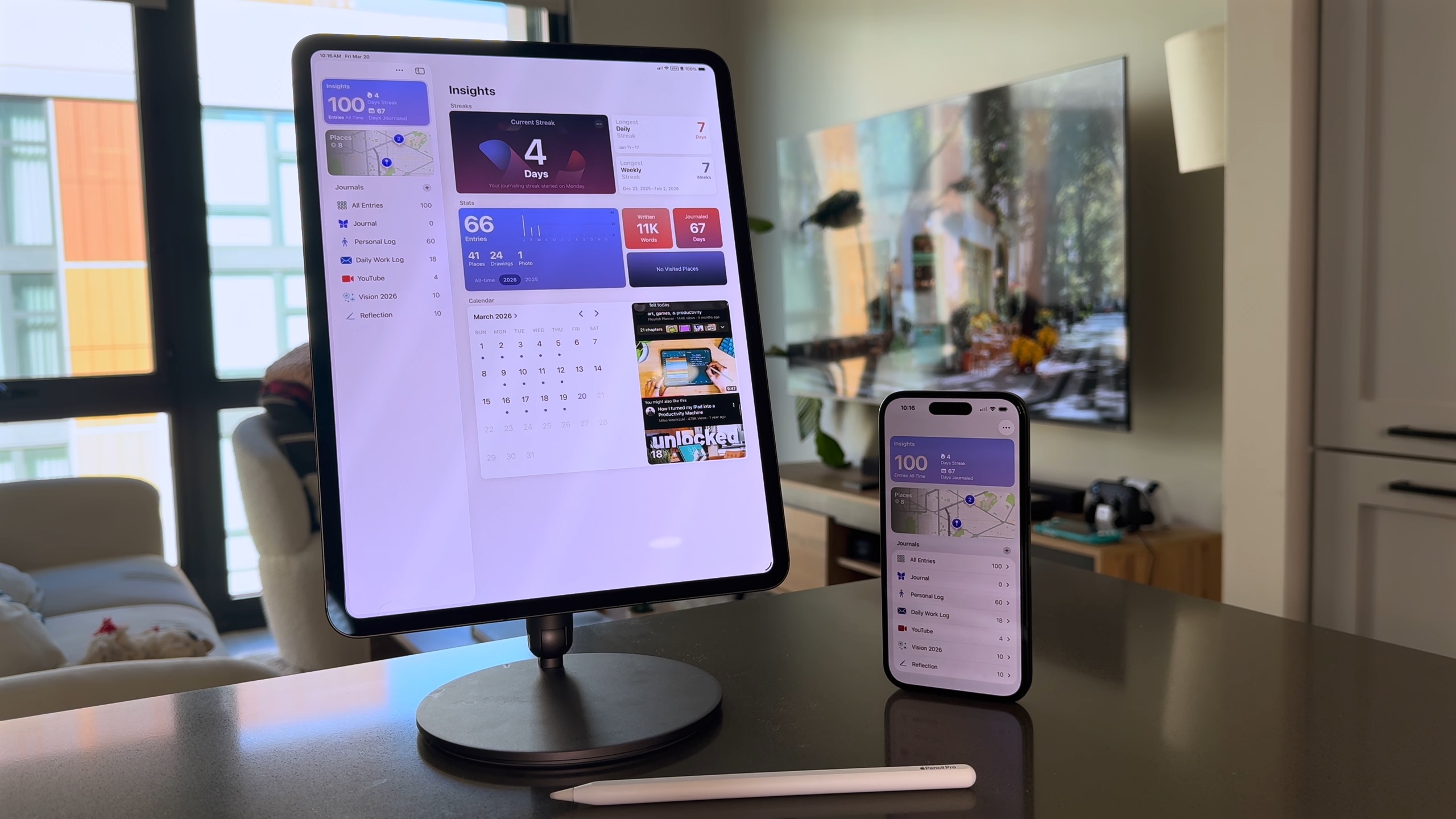 من موقع فون إسلام: جهاز iPad على حامل وجهاز iPhone يعرضان نفس واجهة تطبيق اليوميات آبل؛ وقلم Apple Pencil على الطاولة أمامهما، وفي الخلفية غرفة معيشة عصرية.