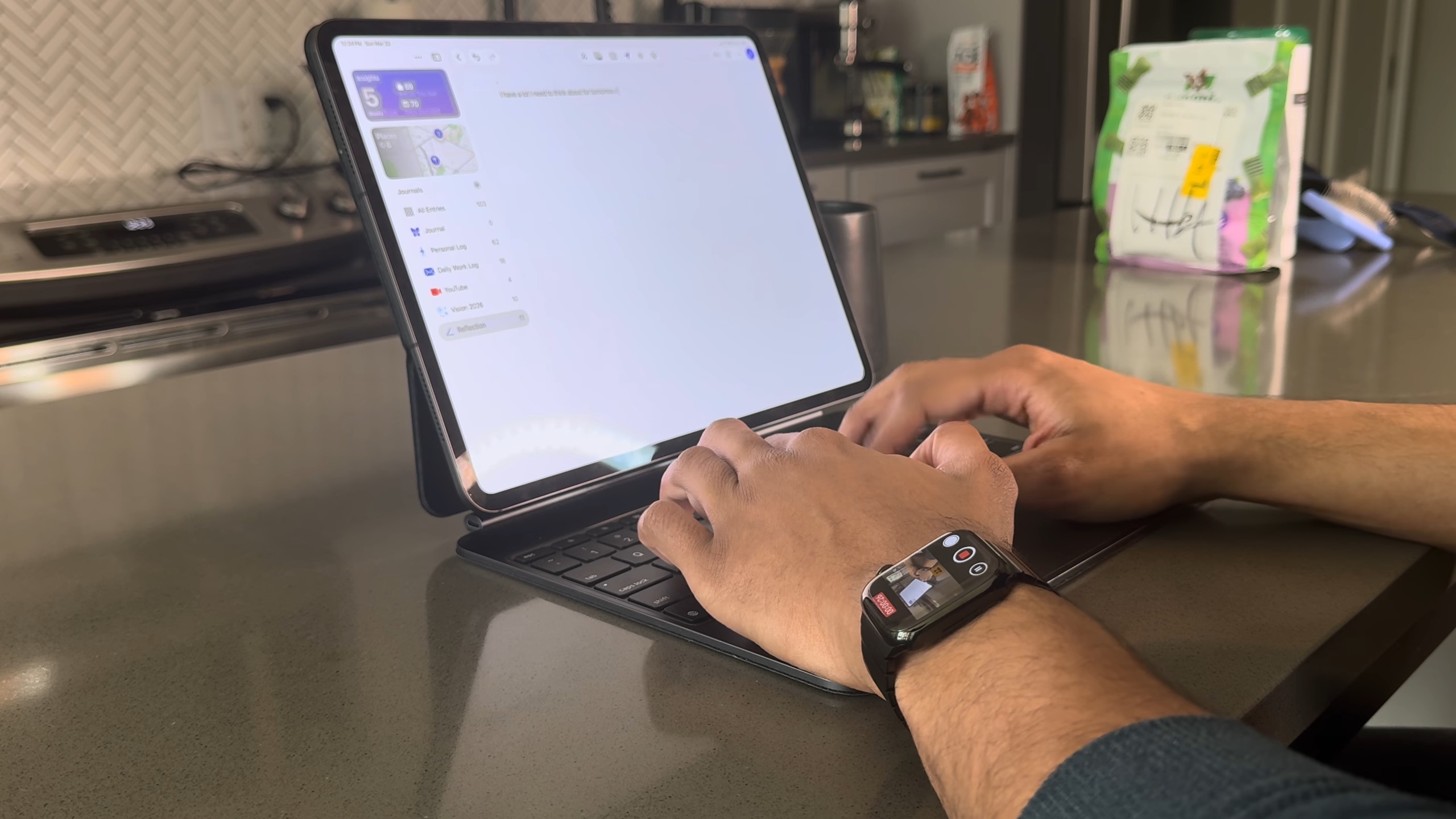 من موقع فون إسلام: شخص يكتب على جهاز لوحي مزود بحافظة لوحة مفاتيح على طاولة المطبخ، وساعة Apple Watch تعرض تطبيق اليوميات آبل آبل مرئية على معصمه.