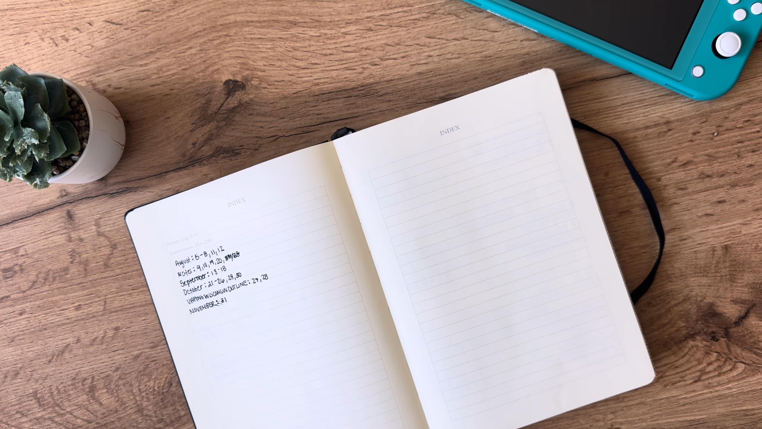 من موقع فون إسلام: دفتر ملاحظات مفتوح على طاولة خشبية يعرض صفحة بها نص مكتوب بخط اليد وصفحة أخرى فارغة. توجد نبتة عصارية صغيرة ونينتندو نينتندو سويتش لايت مخضر، وجهاز آيفون يحمل تطبيق اليوميات آبل في مكان قريب.