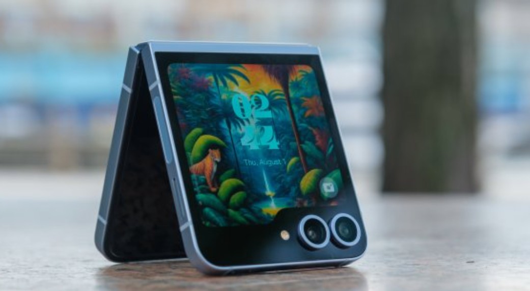 من موقع فون إسلام: هاتف ذكي قابل للطي، يشبه الهاتف الذكي القابل للطي، يشبه الآي القابل للطي مع خلفية ملونة بطابع الغابة مفتوح جزئياً ويقف على سطح خشبي في الهواء الطلق.