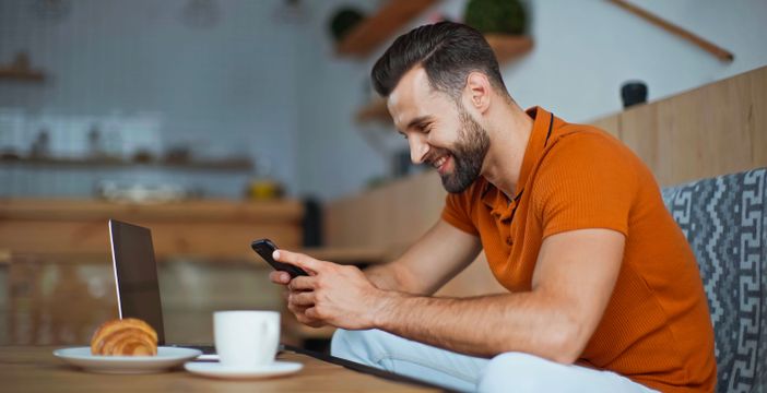 من موقع فون إسلام: رجل يجلس على طاولة مع كمبيوتر محمول وكوب قهوة ومعجنات ويبتسم وهو يتفقد هاتفه بحثًا عن رسائل مؤمنة بواسطة تشفير RCS- İV-فون وأندرويد.