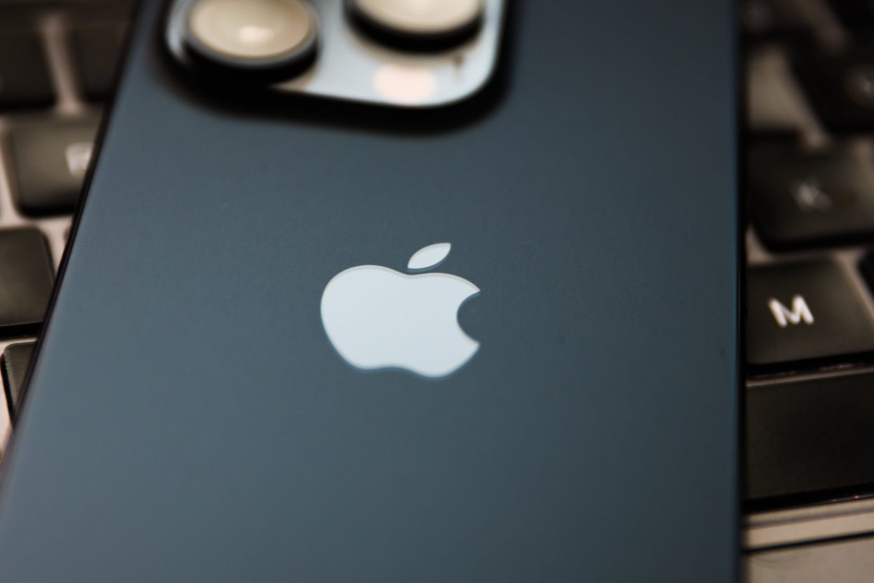 من موقع فون إسلام: لقطة مقرّبة للجزء الخلفي من هاتف آيفون أزرق يحمل شعار Apple، يستند على لوحة مفاتيح كمبيوتر سوداء.