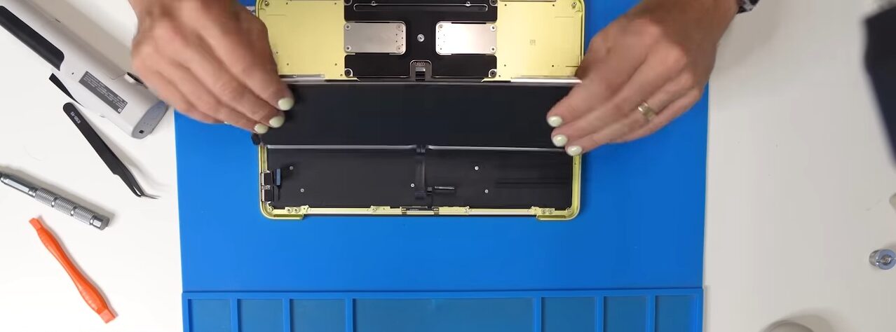 من موقع فون إسلام: شخص يقوم بتفكيك جهاز Nintendo Switch Lite أصفر اللون، مع وضع الأدوات ومكونات الجهاز على حصيرة عمل زرقاء، على غرار إعدادات جهاز MacBook Neo.