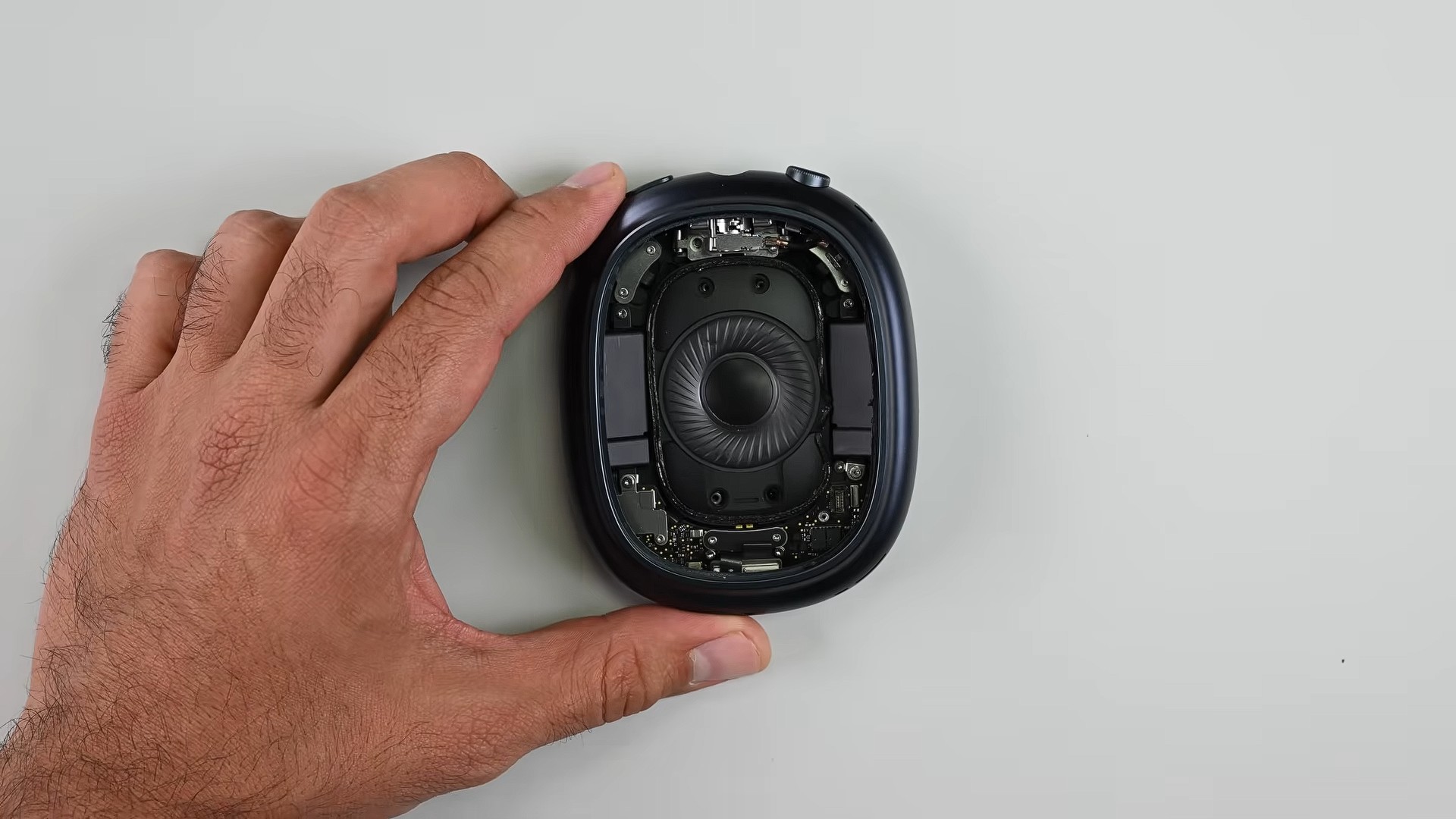 من موقع فون إسلام: يد تحمل سماعة أذن فوق الأذن مع إزالة الغطاء الخارجي، كاشفة عن المكونات الداخلية والدوائر الكهربائية أثناء تفكيك آير-بودز ماكس 2.