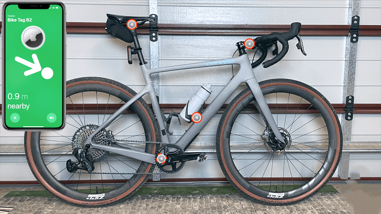 من موقع فون إسلام: توجد دراجة رمادية اللون على الطريق مع نقاط مظللة أمام باب مرآب؛ وتظهر شاشة هاتف داخلية تعرض تطبيق تحديد الموقع آبل للتتبع، مما يشير إلى أن علامة الدراجة على بعد 0.9 متر بالقرب من المكان.