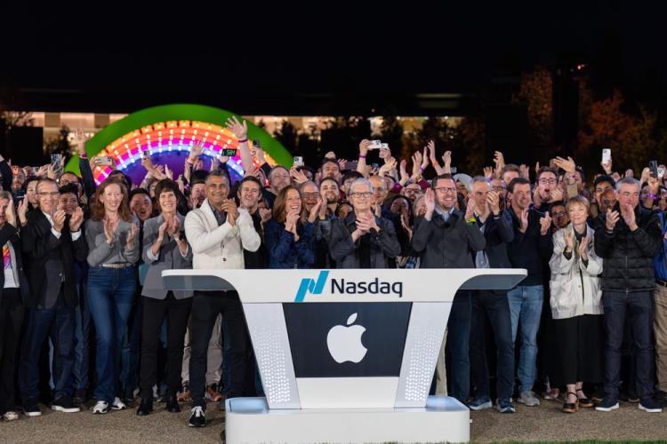 من موقع فون إسلام: مجموعة كبيرة من الناس يقفون خلف منصة ناسداك التي تحمل شعار آبل ويصفقون ويبتسمون في حدث في الهواء الطلق ليلاً خلال احتفال آبل بالذكرى الخمسين.