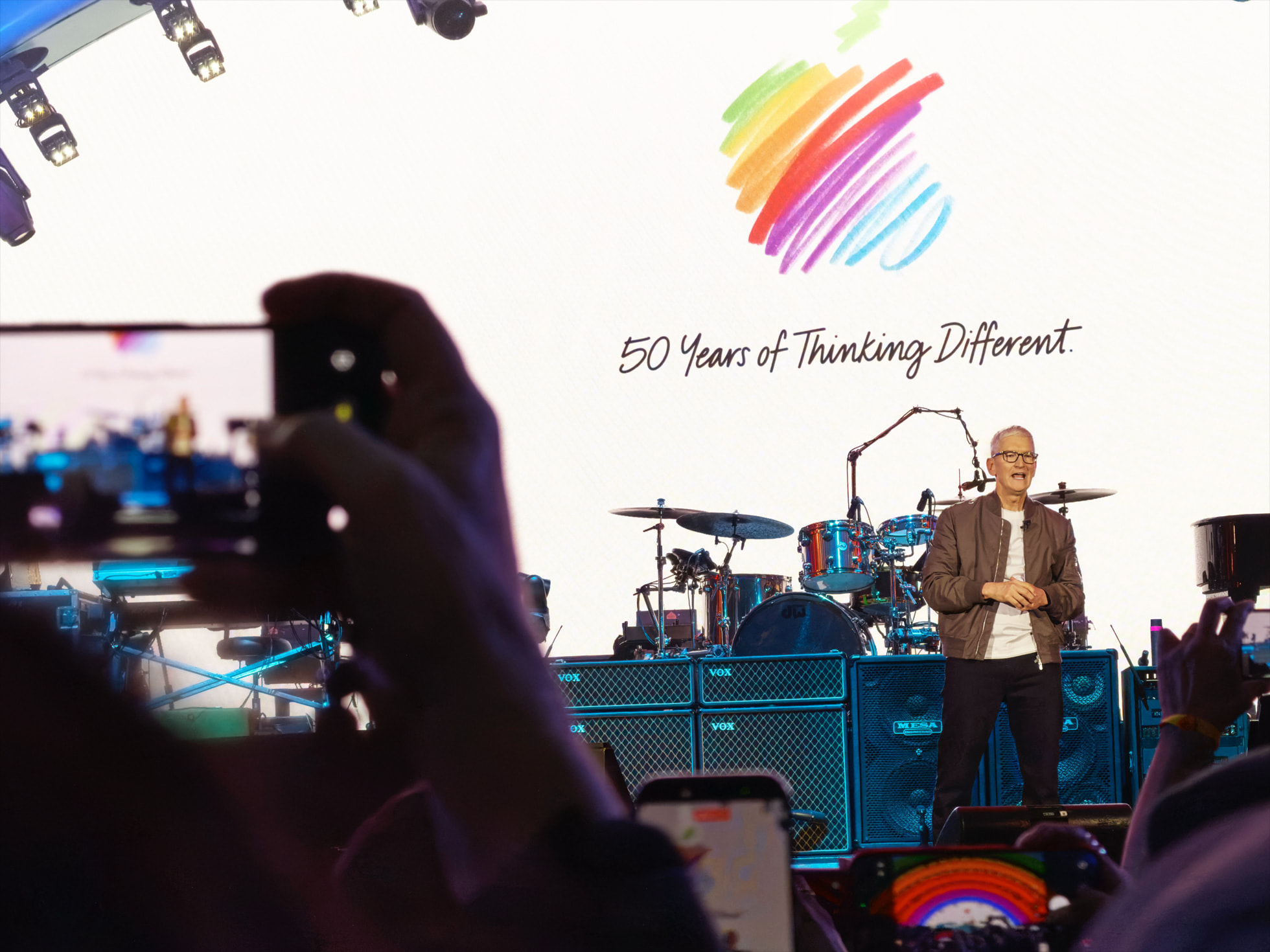 من موقع فون إسلام: شخص يقف على خشبة المسرح أمام طبول وشعار شركة آبل، مع عبارة "50 عامًا من التفكير المختلف" على الشاشة في الخلف، خلال احتفال آبل بالذكرى الخمسين، بينما يلتقط الجمهور الصور التذكارية.