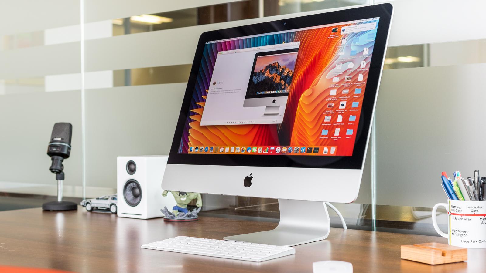 من موقع فون إسلام: جهاز iMac من Apple يعرض شاشة سطح مكتب على مكتب خشبي مع لوحة مفاتيح وماوس ومكبر صوت ومكبر صوت وميكروفون وأقلام وأدوات مكتبية أخرى بالقرب منه.