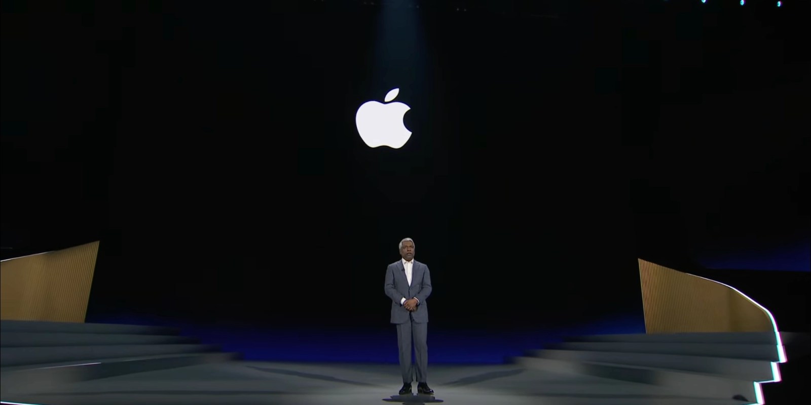 من موقع فون إسلام: يقف رجل يرتدي بدلة على المسرح تحت شعار Apple كبير مضاءً، وتحيط به خطوات وخلفية مظلمة وهو يناقش تطورات مثل سيري وذكاء اصطناعي.