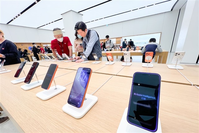 من موقع فون إسلام: تُعرض العديد من الهواتف الذكية على طاولة خشبية في متجر ذي إضاءة ساطعة، حيث يتفحص العملاء الأجهزة - مما يعكس الطلب المتزايد الذي يقوده شحنات آي فون العالمية 2026.