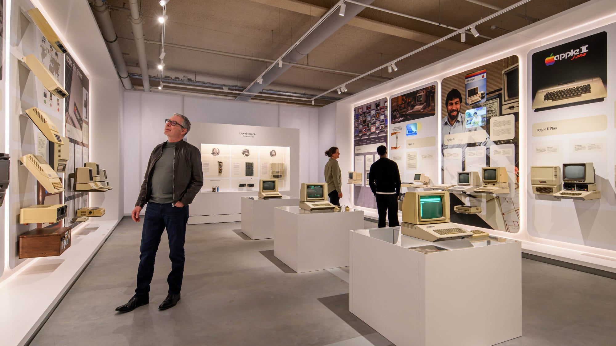 من موقع فون إسلام: ثلاثة أشخاص يشاهدون معروضات الحواسيب والتكنولوجيا القديمة في معرض حديث مضاء جيدًا في متحف آبل في هولندا، محاطًا بشاشات عرض معلوماتية على الجدران.