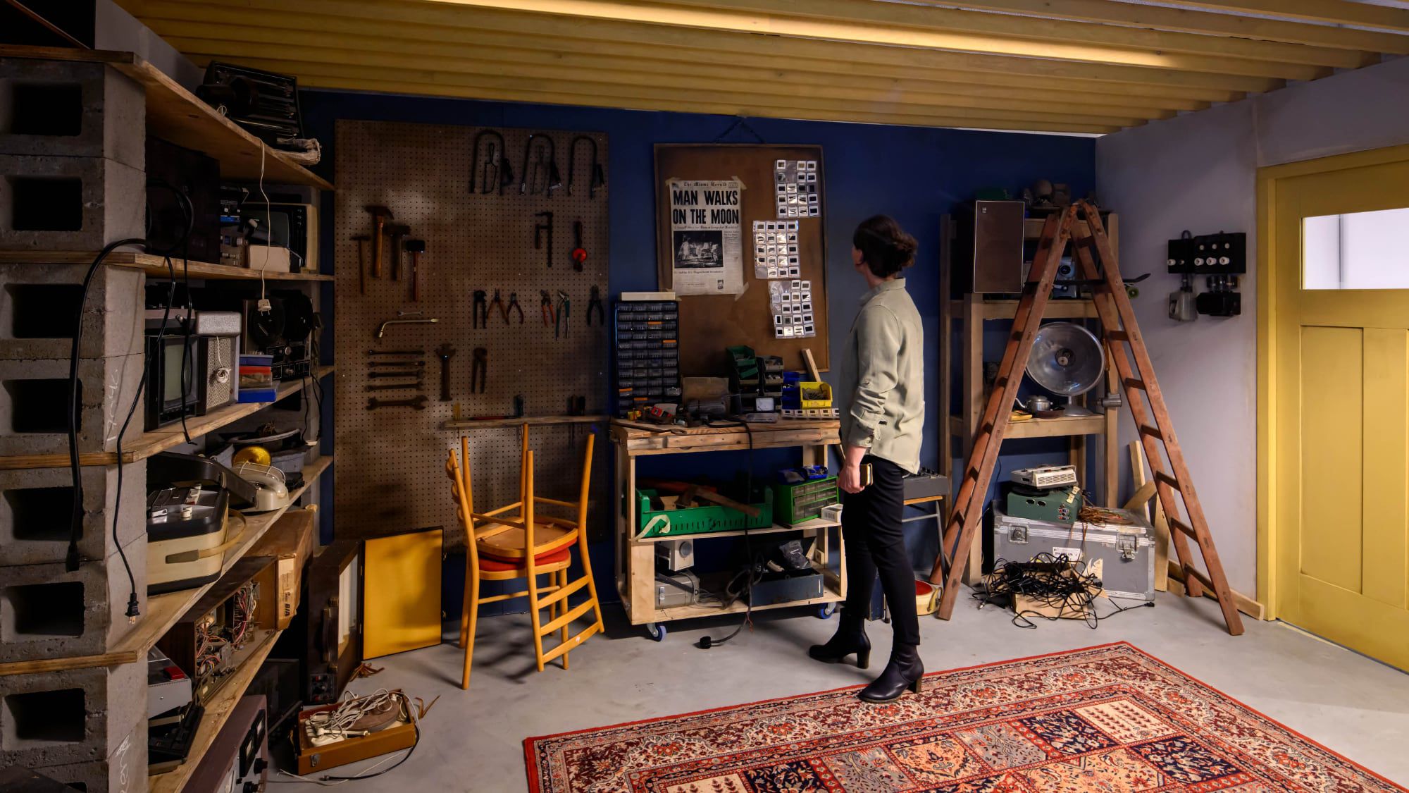 من موقع فون إسلام: يقف شخص في ورشة عمل مرتبة مع أدوات على لوح تعليق ورفوف من المعدات وسجادة على الأرض، ويدرس أغراضًا معلقة على لوحة إعلانات - مثل معرض في متحف في هولندا.