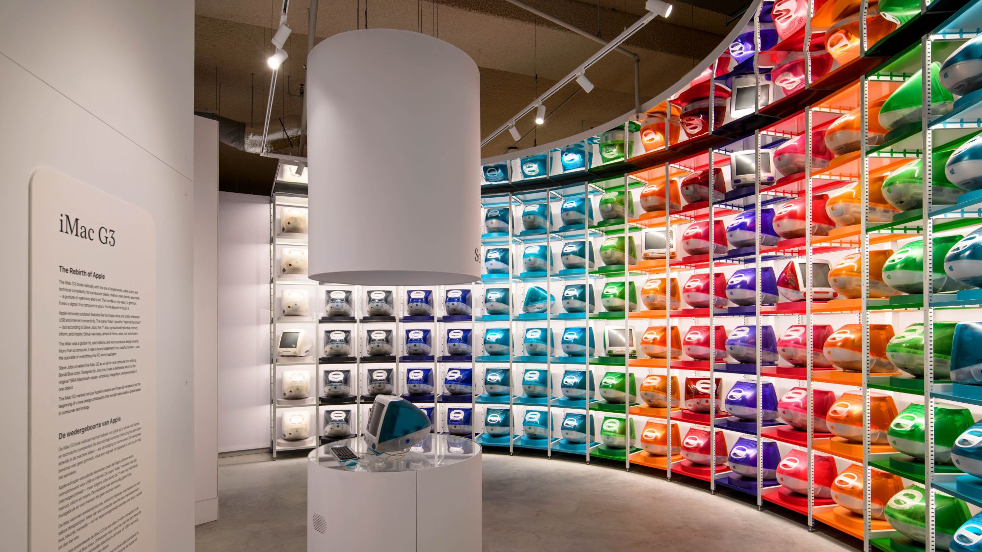 من موقع فون إسلام: شاشة عرض لأجهزة كمبيوتر iMac G3 الملونة مرتبة على أرفف بنمط قوس قزح منحني، مع لوحة إعلامية وحافظة عرض في المقدمة.