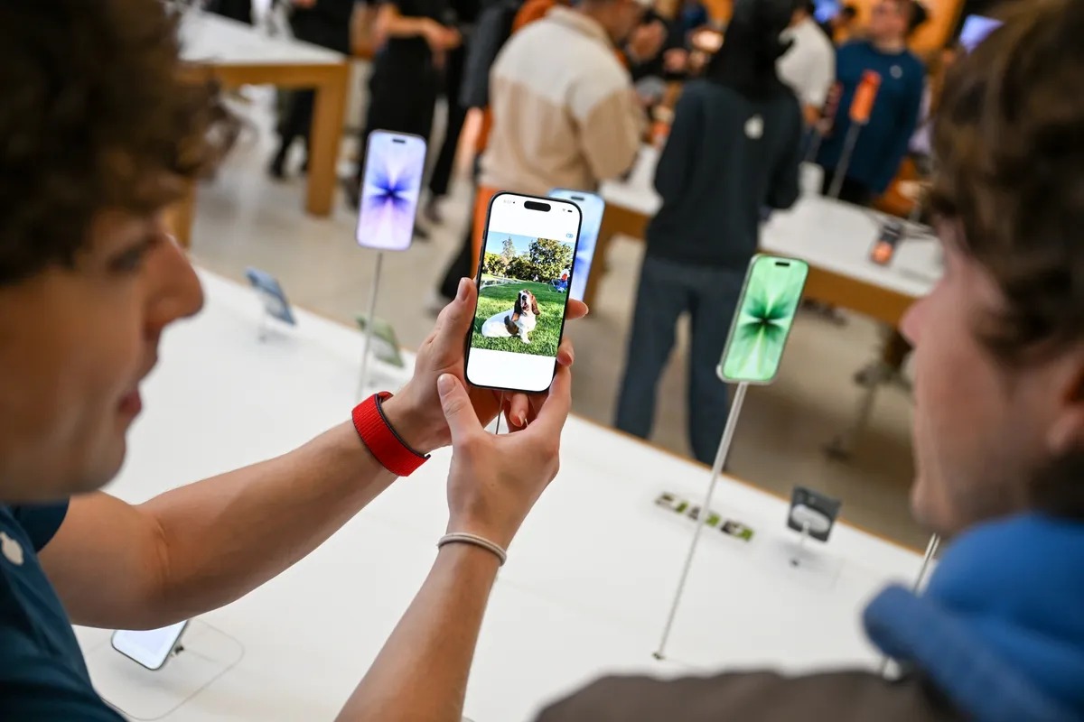 من موقع فون إسلام: شخصان ينظران إلى هاتف ذكي يعرض صورة كلب، داخل متجر إلكترونيات مزدحم، بينما تتداول أخبار الأسبوع تتداول بين الزبائن بين العديد من الهواتف الذكية المعروضة.