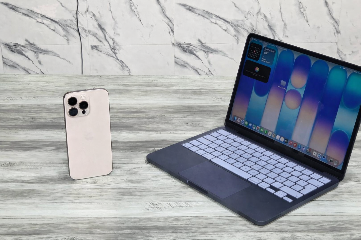 من موقع فون إسلام: يقف هاتف ذكي فضي اللون في وضع مستقيم إلى جانب جهاز كمبيوتر محمول مفتوح يعرض شاشة ملونة، وكلاهما موضوعان على سطح خشبي رمادي اللون مع جدار منقوش بالرخام في الخلفية، مما يسلط الضوء على تجربة ترقية ماك بوك نيو.