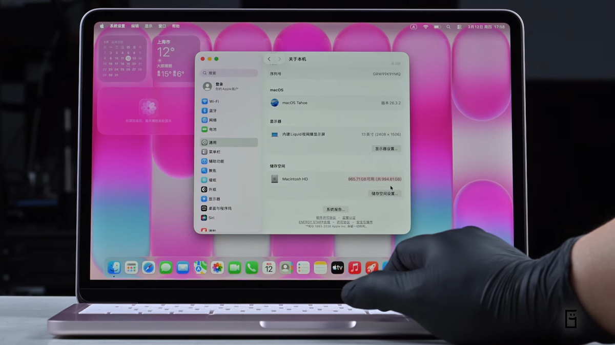 من موقع فون إسلام: شخص يرتدي قفازاً أسود ويشغّل جهاز MacBook Air يعرض نافذة "About This Mac" بخلفية ملونة باللونين الوردي والأبيض، ربما يستعد لترقية ماك بوك نيو.