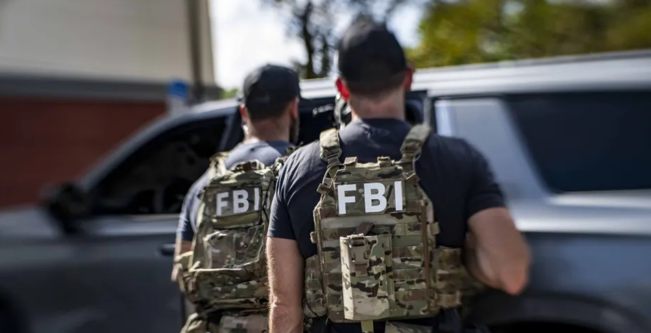 من موقع فون إسلام: يقف عميلان من مكتب التحقيقات الفيدرالي يرتديان معدات تكتيكية بجانب سيارة دفع رباعي سوداء اللون ويواجهان الكاميرا بعيدًا عن الكاميرا، ويحققان في قضية تتعلق بثغرة في ثيفين آيفون ومخاوف بشأن أمان الهاتف.