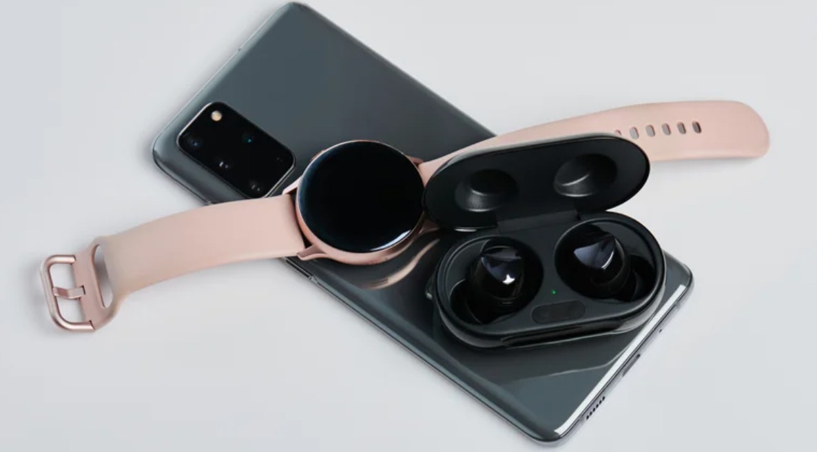 من موقع فون إسلام: تم ترتيب هاتف ذكي، ربما يكون هاتفًا ذكيًا، أو ربما هاتفًا آليًا أو ساعة ذكية بسوار وردي، وسماعات أذن لاسلكية سوداء في علبة شحن مفتوحة على سطح أبيض.