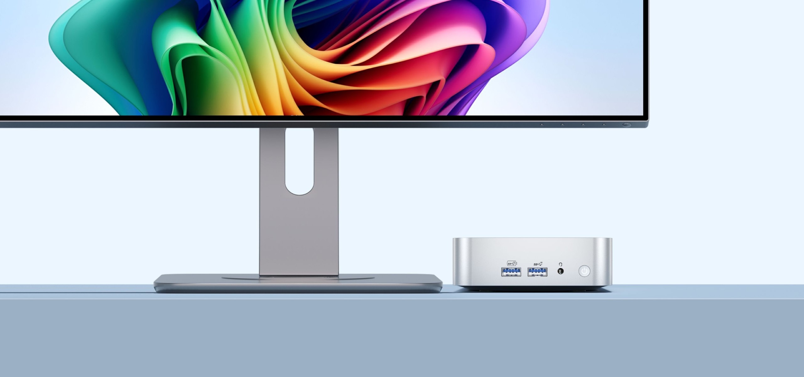 من موقع فون إسلام: شاشة كمبيوتر مزودة بشاشة عرض ملونة تجريدية ملونة تقف بجانب جهاز صغير فضي اللون جهاز GEEKOM A6 كمبيوتر ويندوز على سطح أزرق.