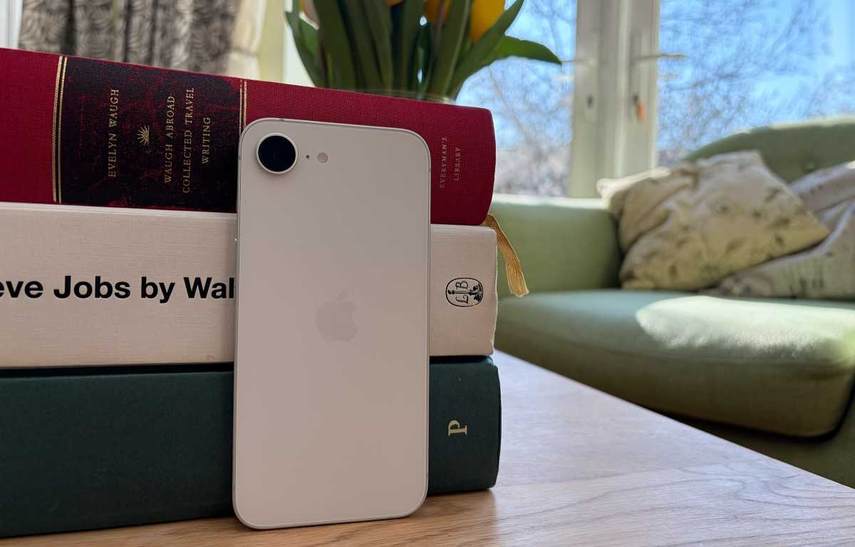 من موقع فون إسلام: يستند هاتف آيفون أبيض على كومة من الكتب على طاولة خشبية، وفي الخلفية أريكة خضراء ونافذة خضراء.