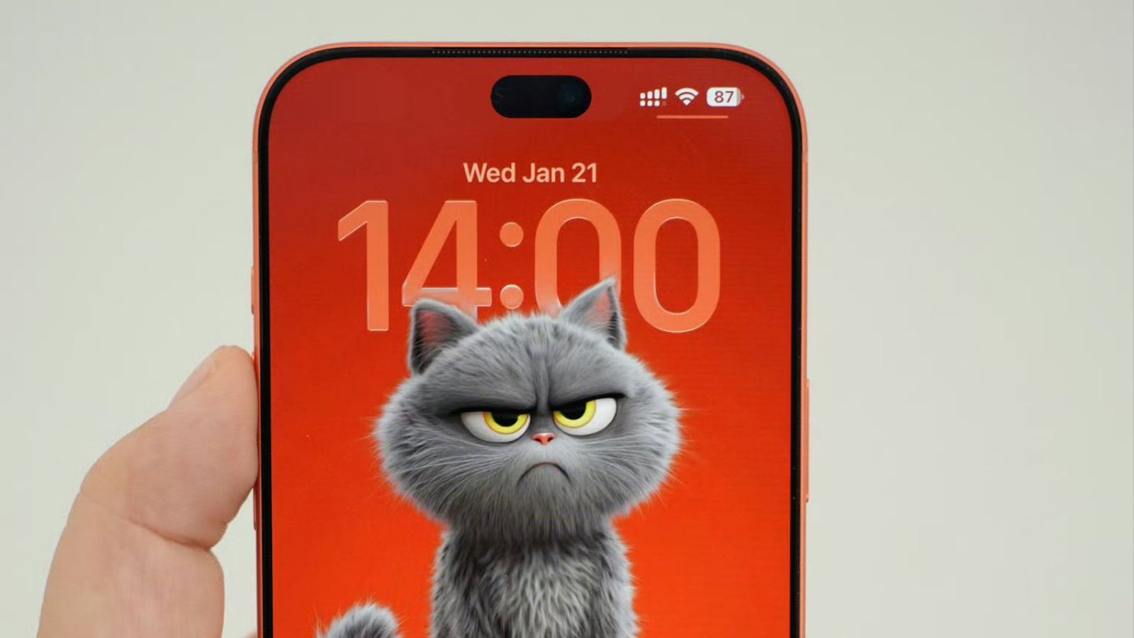 من موقع فون إسلام: يد تحمل هاتفًا ذكيًا يظهر شاشة القفل بخلفية حمراء وساعة كبيرة ورسم توضيحي لقط كرتوني رمادي غاضب - مثالي للبقاء على اطلاع على أخبار التقنية أو ملخص أسبوعي.
