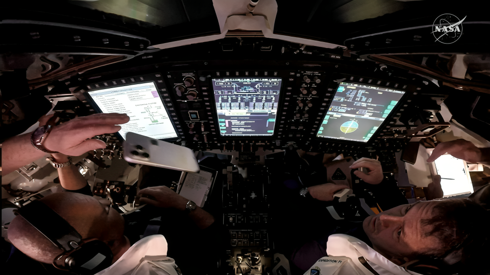 من موقع فون إسلام: يقوم رائدا فضاء داخل قمرة القيادة في المركبة الفضائية بتشغيل أدوات التحكم ومراجعة البيانات على شاشات عرض رقمية كبيرة، مع قوائم مراجعة وأجهزة في اليد - بما في ذلك جهاز آي في الفضاء. يظهر شعار ناسا في الزاوية اليمنى العليا.