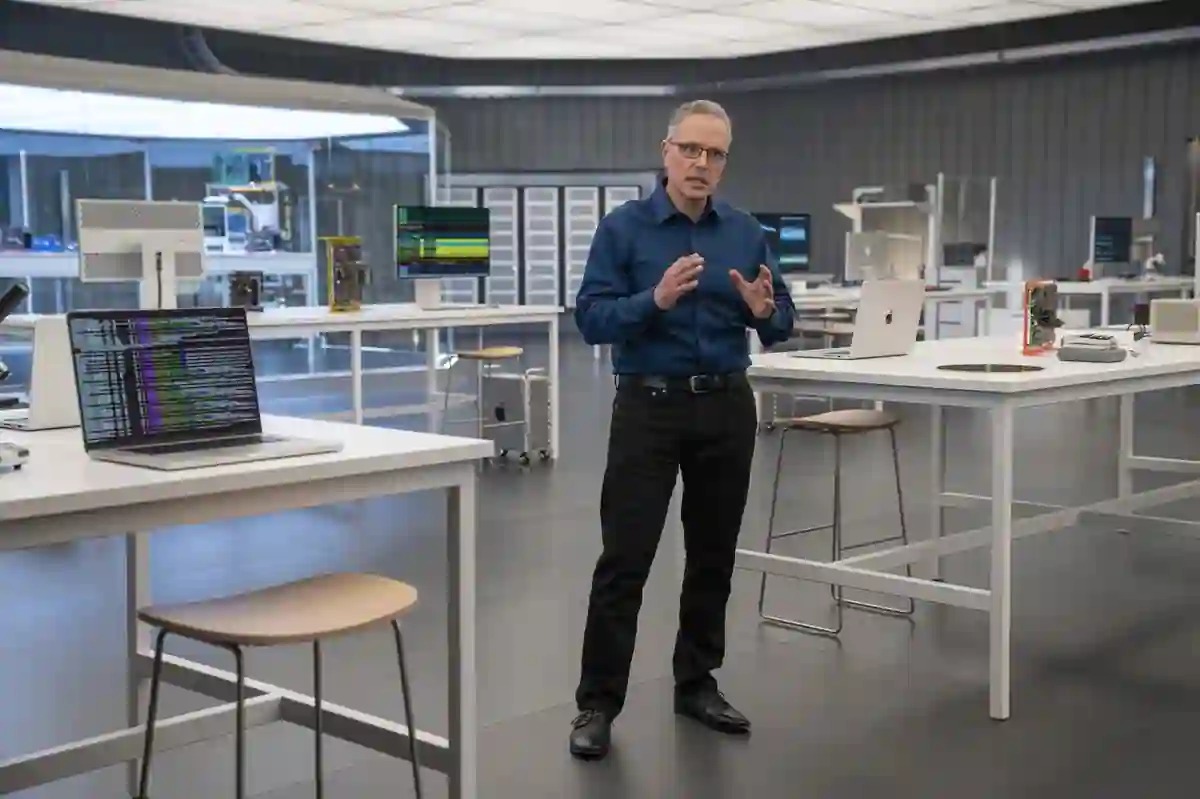 From Phonegram: A man stands in a modern, high-tech lab, speaking while surrounded by computers and electronic equipment on white tables, similar to the innovative environments where Johny Srouji leads technological breakthroughs.