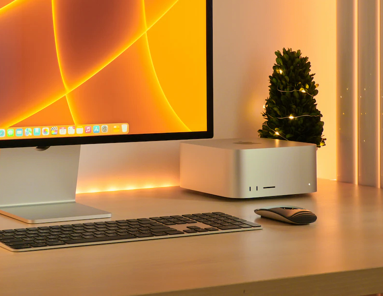 من موقع فون إسلام: إعداد جهاز كمبيوتر مكتبي يضم شاشة ولوحة مفاتيح وماوس وجهاز Mac Studio القادم بجانب نبتة صغيرة مع أضواء خيطية على مكتب خشبي.