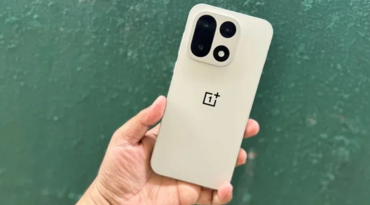 من موقع فون إسلام: يد تحمل هاتفًا ذكيًا أبيض اللون من ون بلس مزودًا بوحدة كاميرا خلفية كبيرة مزودة بطاريات ساتيليكون كربون متطورة، مقابل جدار أخضر اللون.