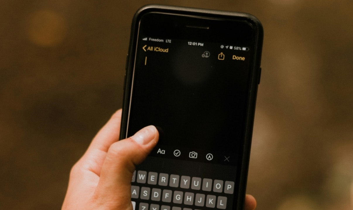 من موقع فون إسلام: يد تحمل هاتفًا ذكيًا مع تطبيق Notes مفتوحًا على ملاحظة فارغة، تظهر لوحة المفاتيح وبعض أيقونات التطبيقات، وهي مثالية لالتقاط أسرار الآي-فون بسرعة وسهولة.