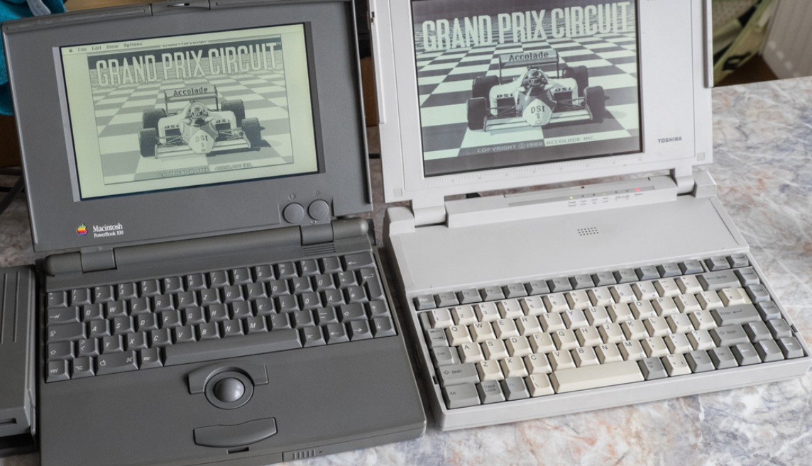 من موقع فون إسلام: تم وضع جهازي كمبيوتر محمول عتيقين، أحدهما رمادي والآخر أبيض، وكلاهما يعرضان نفس شاشة لعبة "حلبة الجائزة الكبرى" جنباً إلى جنب على سطح رخامي.