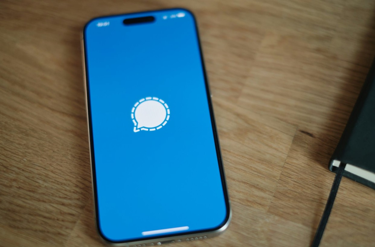 من موقع فون إسلام: هاتف ذكي يعرض شعار تطبيق Signal على شاشته، موضوع على سطح خشبي بجانب دفتر ملاحظات مغلق - مما يسلط الضوء على الحماية من التجسس على الرسائل ثغرات الأمان.