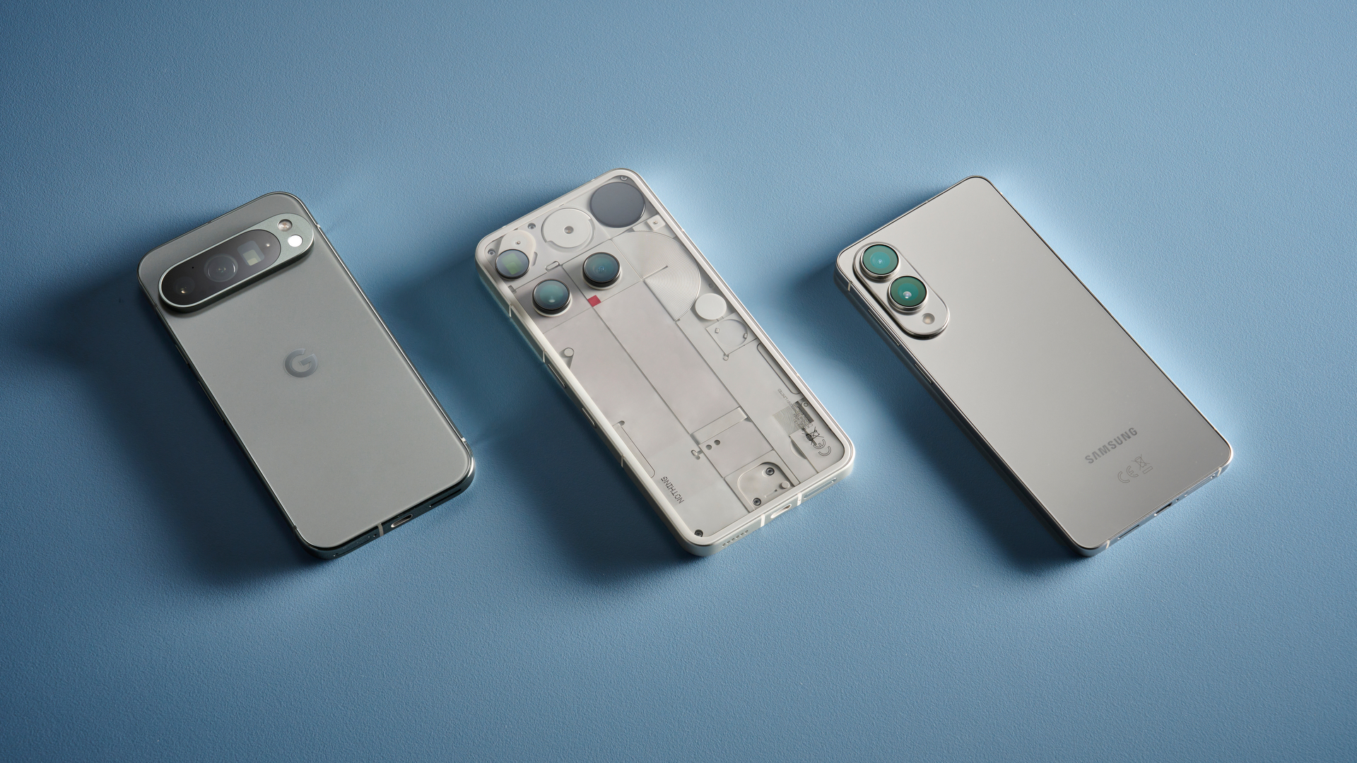 من موقع فون إسلام: وُضعت ثلاثة هواتف ذكية بتصاميم مختلفة على سطح أزرق فاتح، حيث تظهر خلفياتها ووحدات الكاميرا - مما يعكس الابتكار المتوقع مع استمرار شحنات آيفون العالمية 2026 في تشكيل اتجاهات السوق.